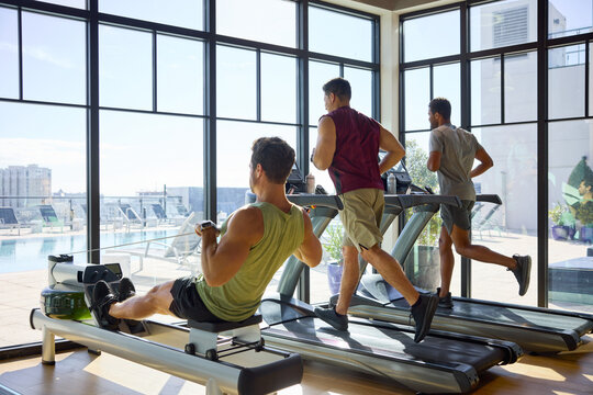 Three men train intensely in a bright gym with city views. They run and row with focus, sweat glistening under. Office in-house gym.