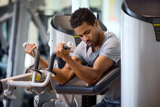 Man focused on strength training at the gym using Preacher Curl Machine. He grips the machine handles, pushing with determination to build muscle. Office in-house gym