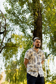 Man in patterned shirt and kippah standing outdoors near a large tree
