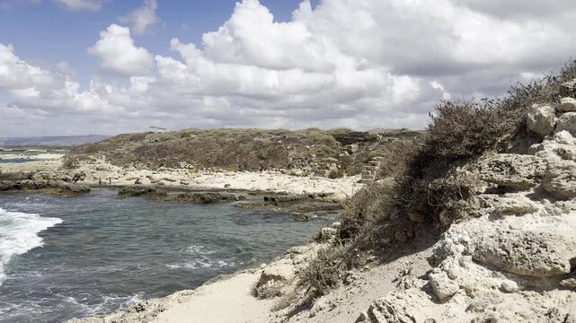 Dor Habonim Beach Nature Reserve, Zichron Yaakov, Israel. View of the Mediterranean coastline from a hill, framed by dry feather grass and ancient stone ruins, with a deserted beach and scattered clou