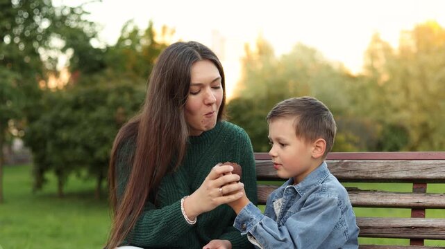 Boy offers chocolate filled cupcake to mother on park bench then takes a bite himself. Slow motion close up shows sharing kindness and family values.