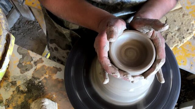 Potter shaping clay bowl on spinning pottery wheel, using clay covered hands to form smooth rounded vessel during traditional ceramic making process