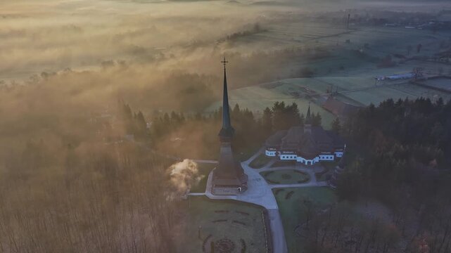 Aerial view of Săp&acirc;nța-Peri Monastery in Maramureș, Romania, emerging through golden morning mist with sunlight spreading across the surrounding rural landscape.