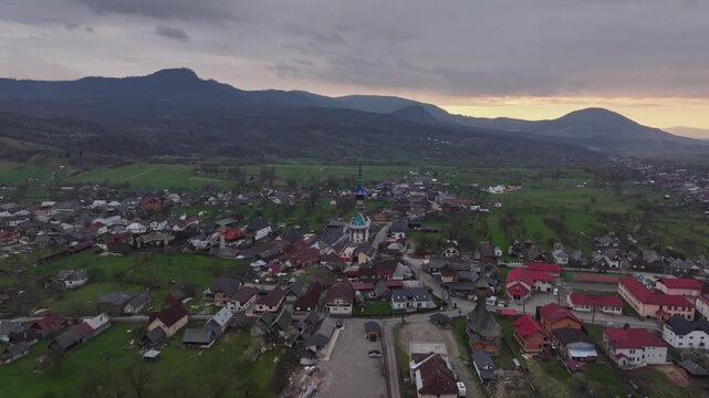 Aerial drone approach toward Săp&acirc;nța village in Maramureș, Romania, with traditional houses, church towers, green fields and mountains under a dramatic cloudy sky.