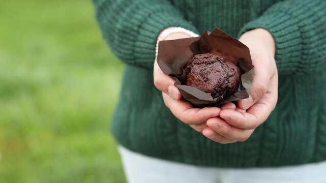 Woman holds chocolate cupcake in paper wrapper close to camera. Visible chocolate pieces reveal rich filling and sweet dessert texture.