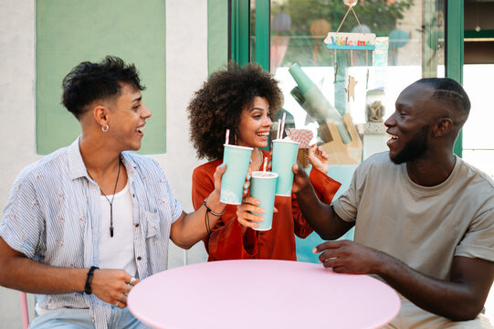 Diverse young friends enjoying contemporary cafe culture, laughing, toasting outside on a summer day