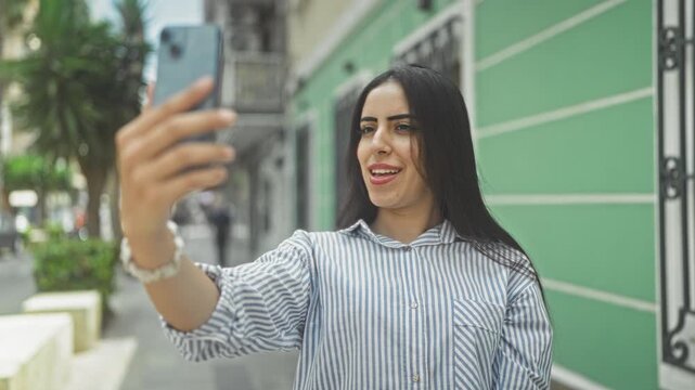 Young arab woman holding smartphone, smiling and waving with hand while taking a selfie on a city street; joyful connection.