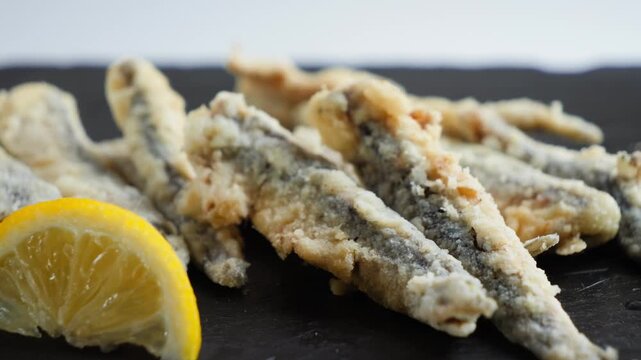 Close-up: a fork picks up a fried European anchovy from the plate; alongside are crispy fish and a lemon wedge.