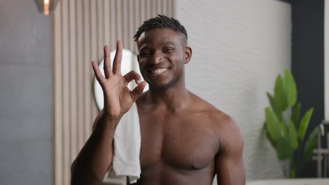 Happy African American muscular man with towel in bathroom showing okay sign smiling looking at camera after morning shower hygiene bath hydration refreshment male guy doing skincare beauty indoors
