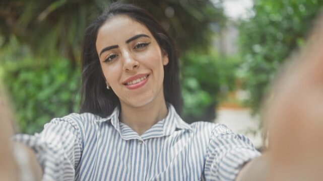 Woman taking a selfie showing face with extended arm on street near green plants and foliage; self expression authentic confidence.