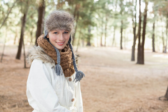 Woman standing left of center in pine clearing, wearing fur trapper hat, knitted scarf, white coat