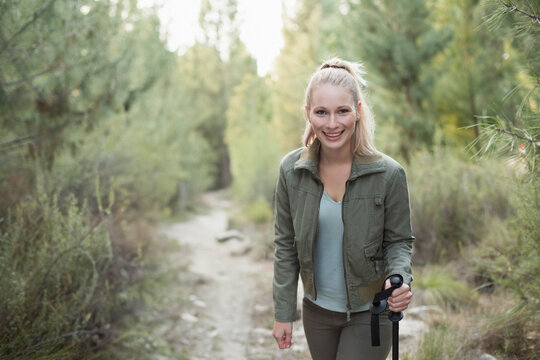 Female hiker standing on dirt trail in forest, holding trekking pole and wearing olive jacket