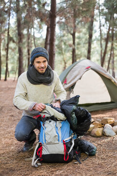 Man wearing beanie kneeling and packing hiking pack at pine campsite, stuffing sleeping bag
