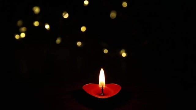 A red heart-shaped candle on a black background with golden bokeh particles flowing down throughout the video, the flame flickers and fades in the dark