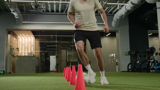 Caucasian footballer man player running with ball indoor gym soccer athlete male sportsman training fast dribbling between cones feint moves dynamic motion legs football workout match speed exercise