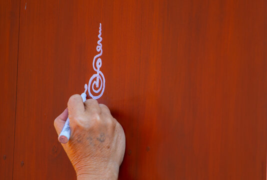Close-up of an adult hand using a white marker to draw a decorative Thai-inspired symbol on a warm wooden surface, with plenty of copy space. Concept of traditional art, creativity, craftsmanship, cul