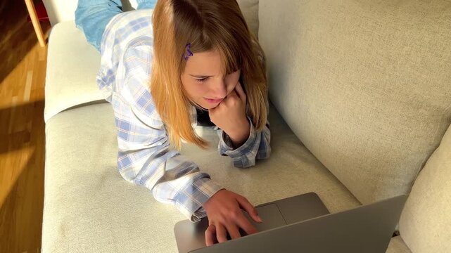Top view of a young woman lying on a light couch and using a laptop, modern indoor leisure, cozy remote studying, casual lifestyle moment.