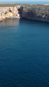 High-angle view of coastal inlet with calm rippling sea, sunlit limestone cliffs, and sheltered cove at shoreline