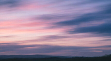 Fototapeta premium Soft pastel sunset sky with wispy clouds in pink and purple hues over a silhouetted landscape, serene atmosphere