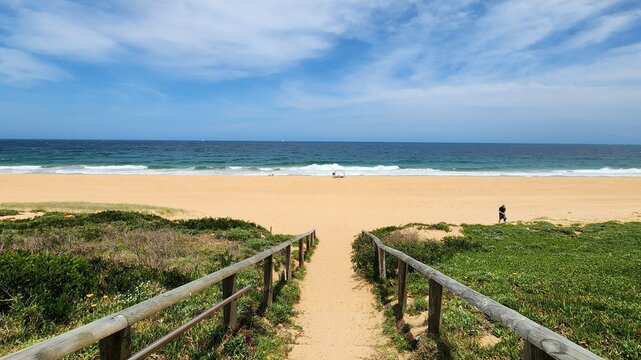 Sydney, Australia - Northern Beaches sandy pathway with wooden railings leading to wide golden beach and blue ocean waves under partly cloudy sky creating a serene coastal landscape.
