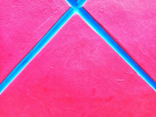 geometric X-shaped pattern in blue placed on a bright red surface. The blue lines meet at the center, creating a simple, symmetrical, and visually striking composition. The red surface has a subtle te © Slamet