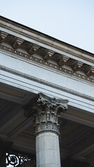 Vertical close-up view of classical building facade featuring ornate Corinthian column capital, detailed carved frieze, and sculptural relief medallion. © Вита Курило