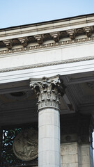 Vertical close-up view of classical building facade featuring ornate Corinthian column capital, detailed carved frieze, and sculptural relief medallion. © Вита Курило