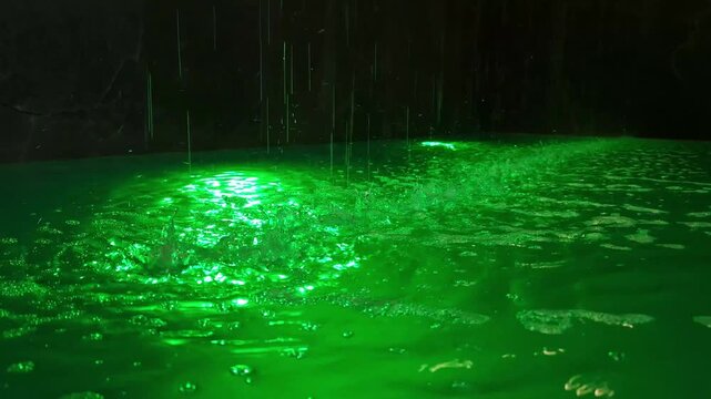 Heavy water droplets fall continuously onto the surface of a brightly illuminated green pool. Ripples, bubbling spray, and glowing reflections form energetic patterns in dark surroundings.