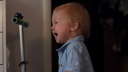 A young toddler with blonde hair crying while standing indoors in a casual blue button-up shirt © Тарас Белецкий
