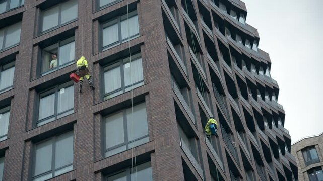 Rope access workers suspended on tall brick building facade beside rows of windows with bright safety gear, tool containers, and geometric architecture under pale overcast city light