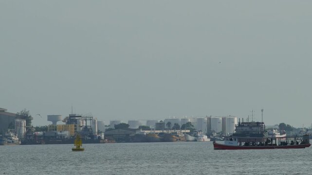 Red wooden passenger ferry boat crossing wide river with passengers, hazy harbor skyline of cargo ships and white oil storage tanks in distance, calm water