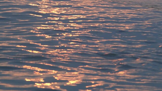 Golden sunset light reflecting and sparkling on gentle blue water ripples, warm shimmering path across calm river surface, tranquil evening footage