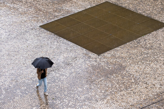 Person walking with an umbrella across patterned paving seen from above creating a minimalist urban composition of geometry movement and scale