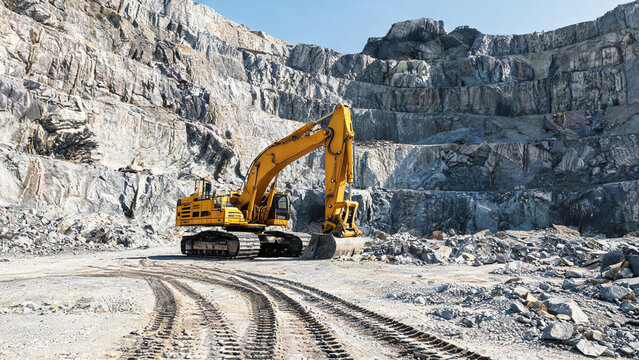 A yellow tracked excavator works at an open-pit stone quarry, with rocky terraces and dirt tracks.