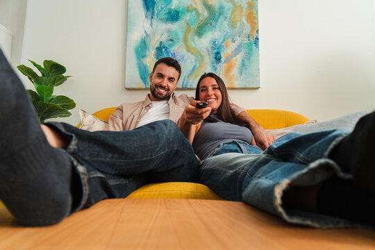 Two people lounge on a yellow sofa, holding a remote and watching television with a playful, relaxed mood. The scene emphasizes togetherness, comfort, and shared entertainment in a cozy living space.