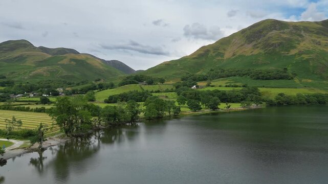 Calming waters reflect rolling green hills under soft clouds. Quiet village nestles beside the lake, peaceful. Buttermere and Grassmere are lakes located in the Lake District National Park in Cumbria