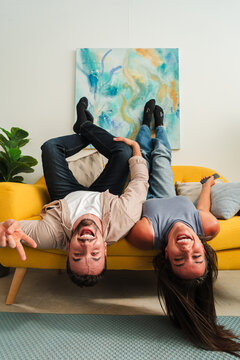 A candid shot of a couple hanging upside down from a sofa, sharing wide smiles and playful energy. The moment exudes joy, balance, and carefree humor in a cozy living room. Fresh energy fills scene.