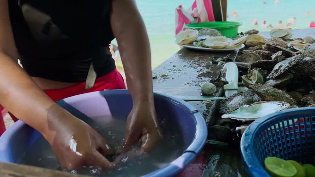Person preparing fresh oysters at outdoor seafood stall, handling shellfish beside a blue water basin with open shells and seafood displayed on table near the sea