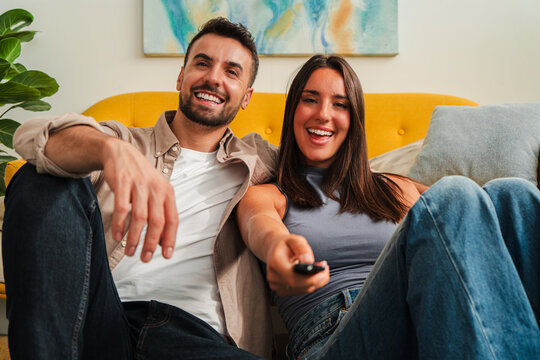 A cheerful couple sits close on a bright yellow couch, smiling toward the camera while they use a handheld remote to control a television This moment conveys relaxed home entertainment and warm mood
