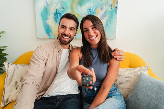 A warm portrait shows a smiling couple on a bright yellow sofa, one pointing a TV remote toward the viewer as they share a relaxed, intimate moment during a casual night in.
