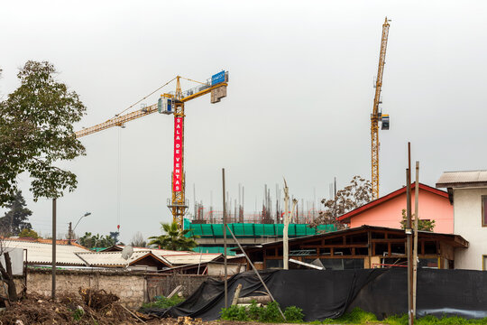 Two large tower cranes rise over a construction site in Santiago, Chile, contrasting with nearby residential homes and an overcast sky