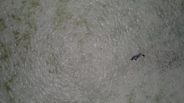 Drone high-angle overhead shot two fishermen standing in shallow, crystal-clear tropical sea water while fly fishing. Clear water ripples texture showing sandy seabed underneath. Flat color profile