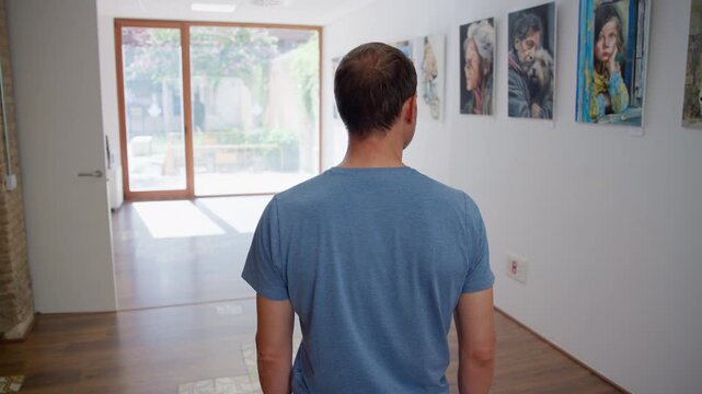Camera follows a man from behind as he walks down a gallery corridor lined with framed artwork, Slow Motion.