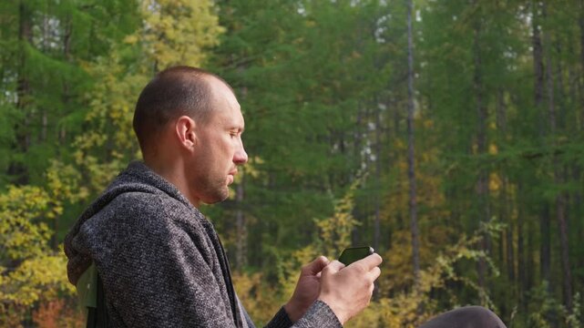 A man in a gray hooded sweater sits in an autumn forest, intently using his black smartphone with minimal finger movement, surrounded by trees with green and yellow leaves under overcast lighting.
