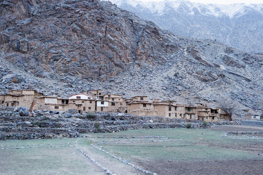Traditional stone and mud houses in rural mountain village nestled beneath steep rocky scree slope, Remote alpine village with vernacular mud brick masonry homes below rocky mountain cliff