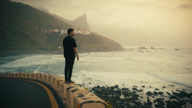 A man standing on a coastal barrier overlooking the village of Benijo in the Anaga mountain range, Tenerife, Spain, in a cinematic golden sunset shot.
