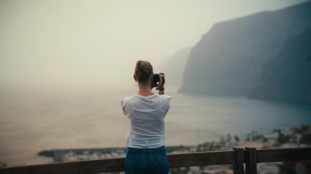 Woman photographing the hazy sea cliffs of Los Gigantes, Tenerife, from a coastal viewpoint under soft, calima-diffused light.