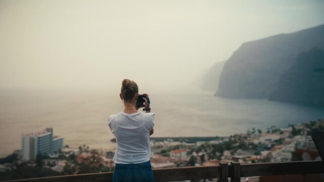 Woman photographing the coastal town and hazy sea cliffs of Los Gigantes, Tenerife, from an elevated viewpoint under soft, calima-diffused light.