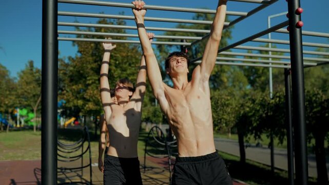 Shirtless Generation Z boy doing dynamic pull ups on monkey bars at the park.