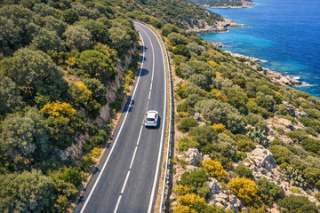 Obraz premium Sunny day, winding road, car on right side, green foliage, blue water, Mediterranean-style road, travel, outdoor adventure